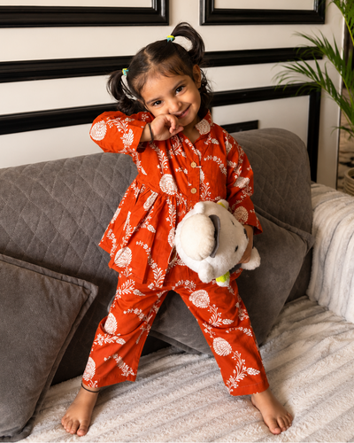 Child in rust Dori Co-ord dress from Aruvo with white patterns standing on a couch holding a teddy bear, with a plant and framed pictures in the background.