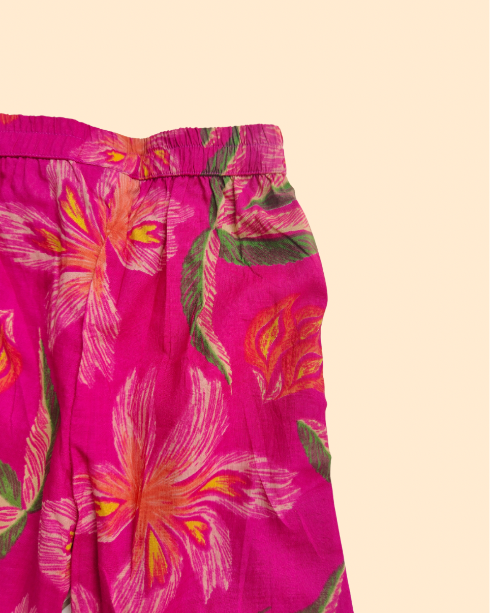 Bright pink floral printed pant with a gathered elastic waistband, featuring large flowers in pink, orange, and yellow with green leaves, shown against a solid pale beige background from Aruvo