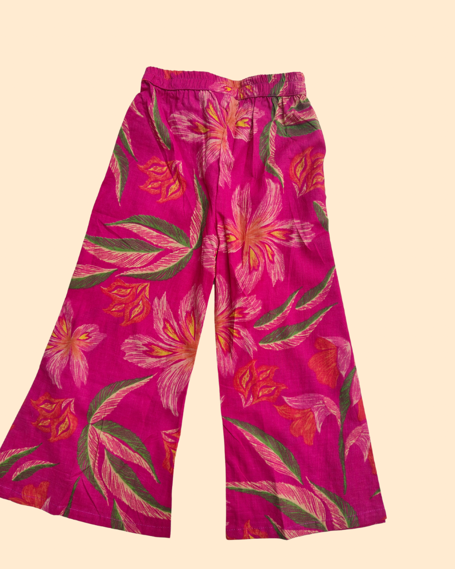 Bright pink wide-leg pants with a tropical floral and leaf print in shades of green, orange, and white, displayed flat against a light beige background.
