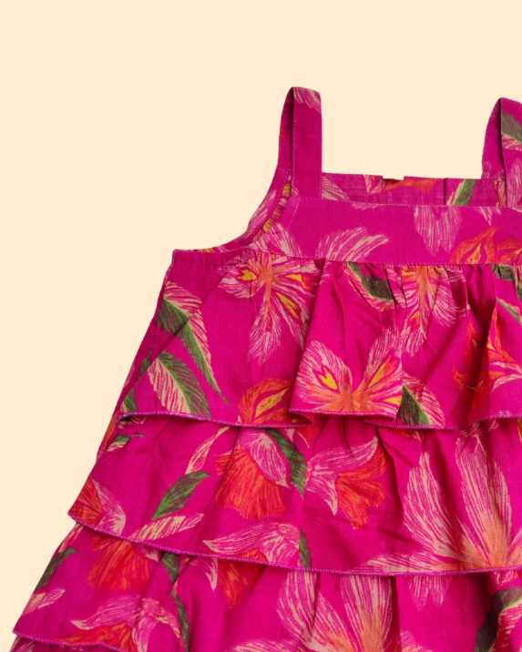 A tiered sleeveless dress for toddlers or girls, featuring a vibrant magenta pink base with a tropical floral print in orange, red, and green, displayed against a solid pale beige background.