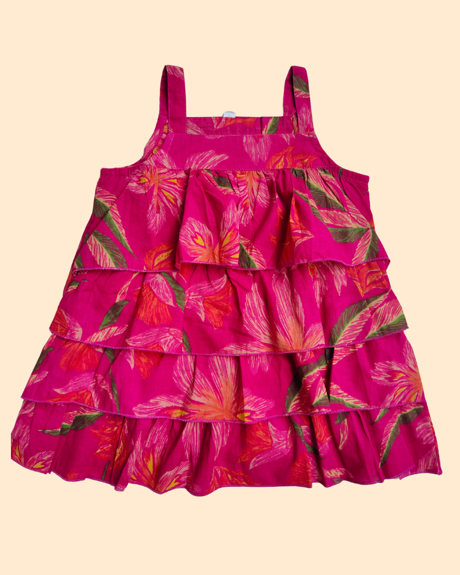 A tiered ruffle babydoll dress in vibrant magenta pink with a tropical floral print in orange, yellow, and green, shown against a solid pale beige background from Aruvo