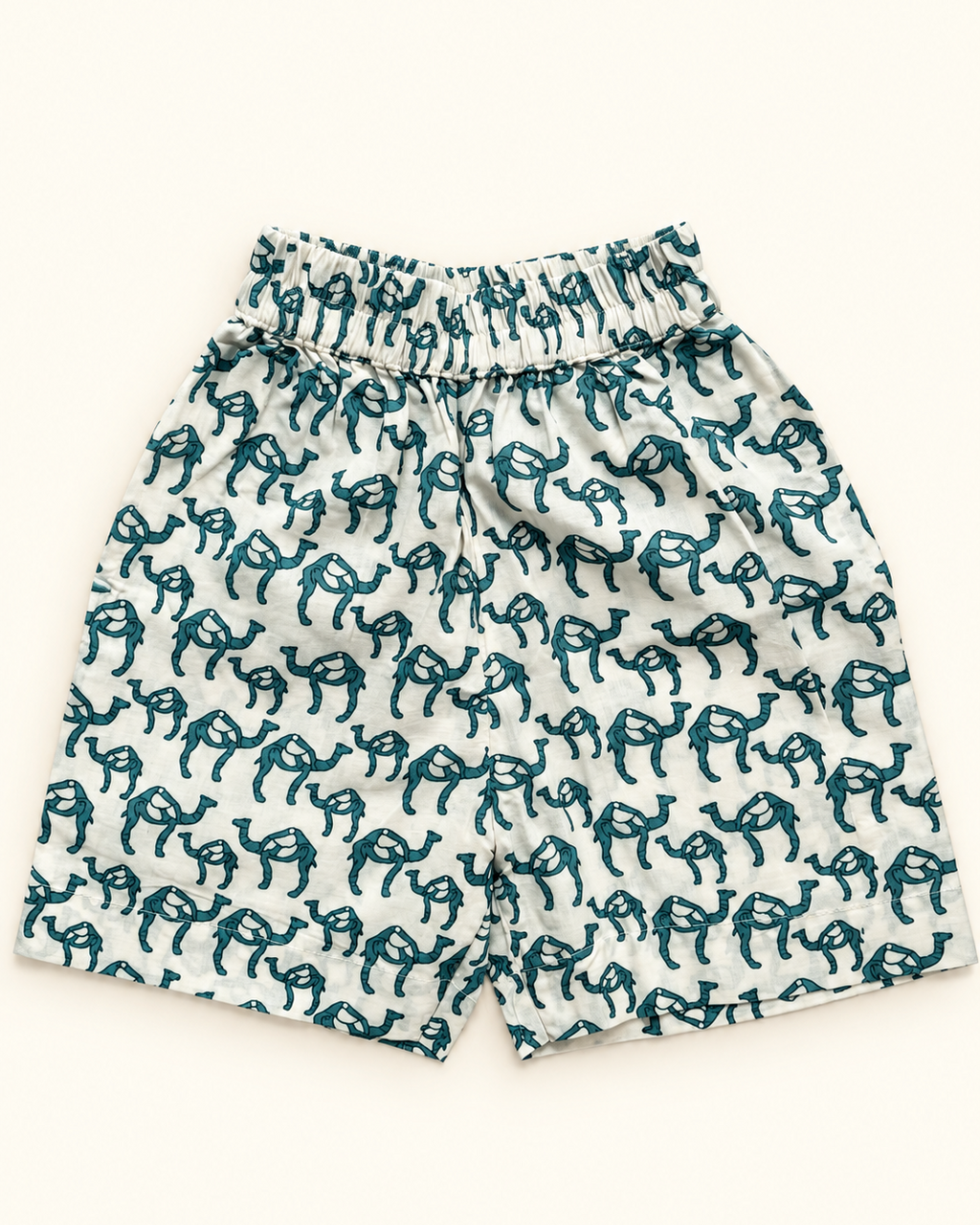 A pair of white shorts with an all-over dark teal camel print, featuring an elastic waistband, laid flat against a light cream background.