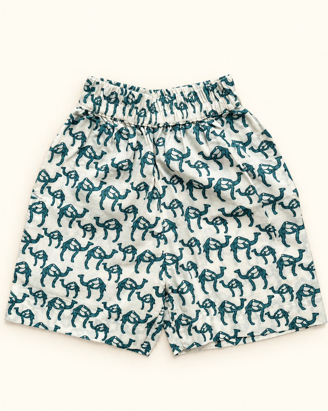 A pair of white shorts with an all-over dark teal camel print, featuring an elastic waistband, laid flat against a light cream background.