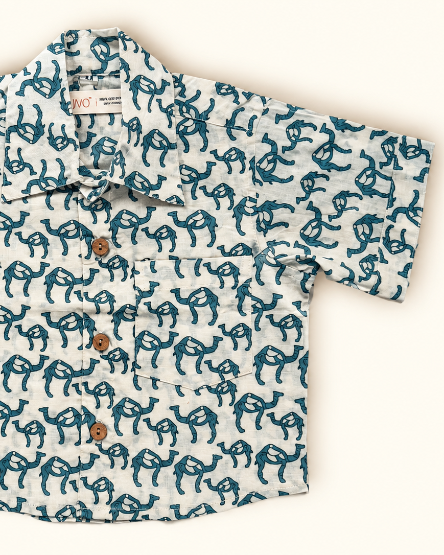 A children's long-sleeve shirt with an all over teal and white abstract pattern, featuring two front pockets with brown buttons, laid flat on a light cream background.