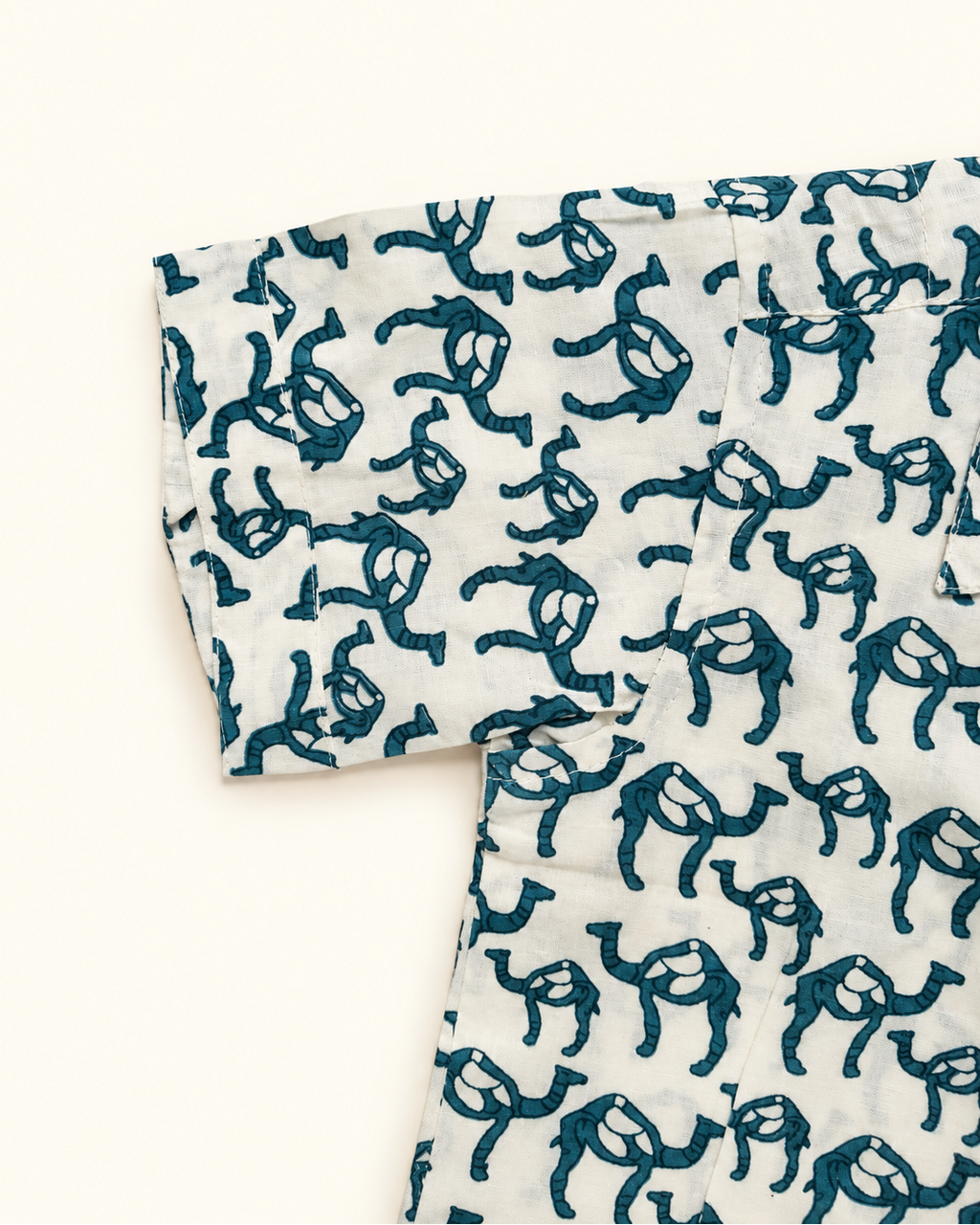 A close-up of a white fabric garment featuring an all-over dark teal block-print camel pattern, laid flat against a solid off-white background.
