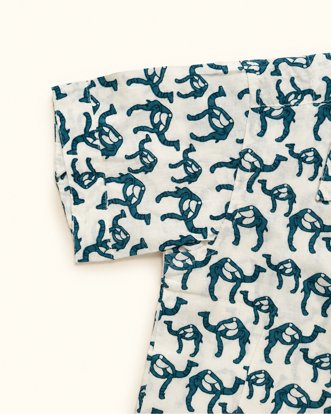 A close-up of a white fabric garment featuring an all-over dark teal block-print camel pattern, laid flat against a solid off-white background.