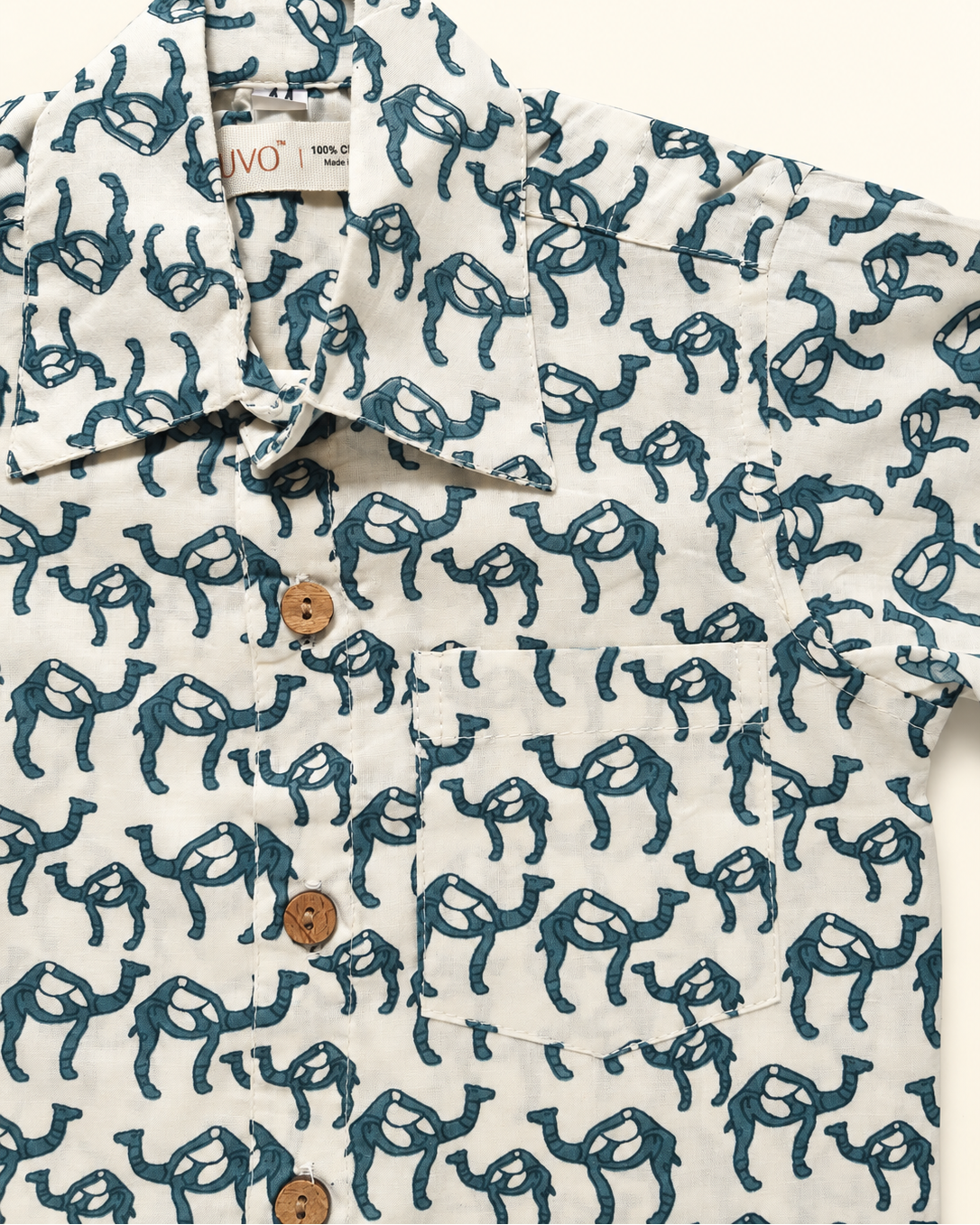 A close-up of a short-sleeve button-up shirt by ARUVO, featuring an all-over camel print in dark teal on a warm off-white background. The shirt has a pointed collar, a chest pocket, and brown wooden buttons.