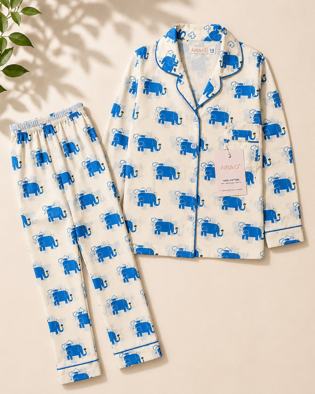 A two-piece pajama set laid flat on a warm beige background, featuring a white long-sleeve top and matching pants with an all-over blue elephant print and blue piping. A brand tag for 'ARUVO' is visible on the shirt.