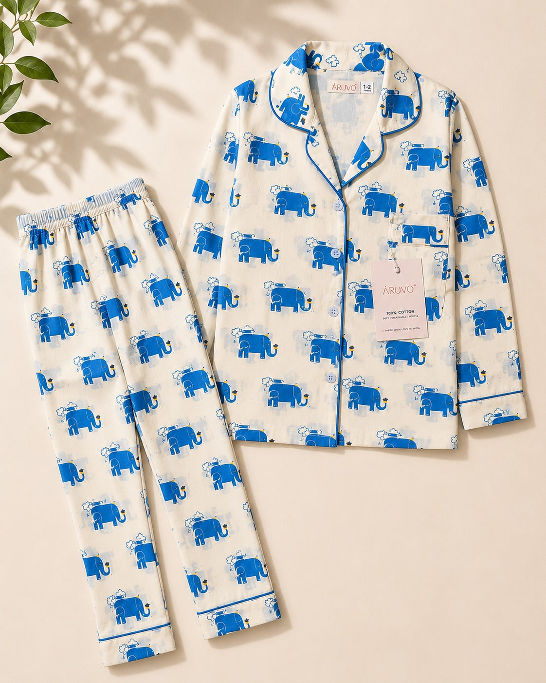 A two-piece pajama set laid flat on a warm beige background, featuring a white long-sleeve top and matching pants with an all-over blue elephant print and blue piping. A brand tag for 'ARUVO' is visible on the shirt.
