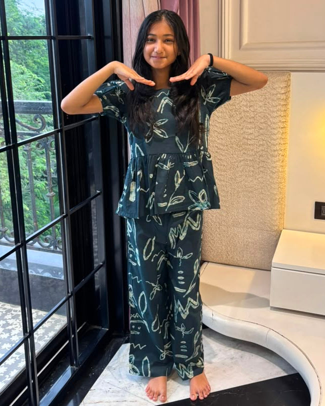 A girl is wearing a peplum styled top with matching pant in navy blue color from brand Aruvo, standing near a glass door smiling