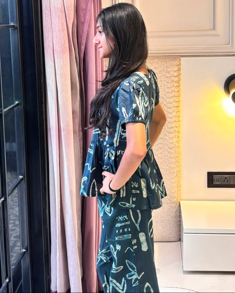 A girl is wearing a peplum styled top with matching pant in navy blue color from brand Aruvo, standing near a glass door smiling