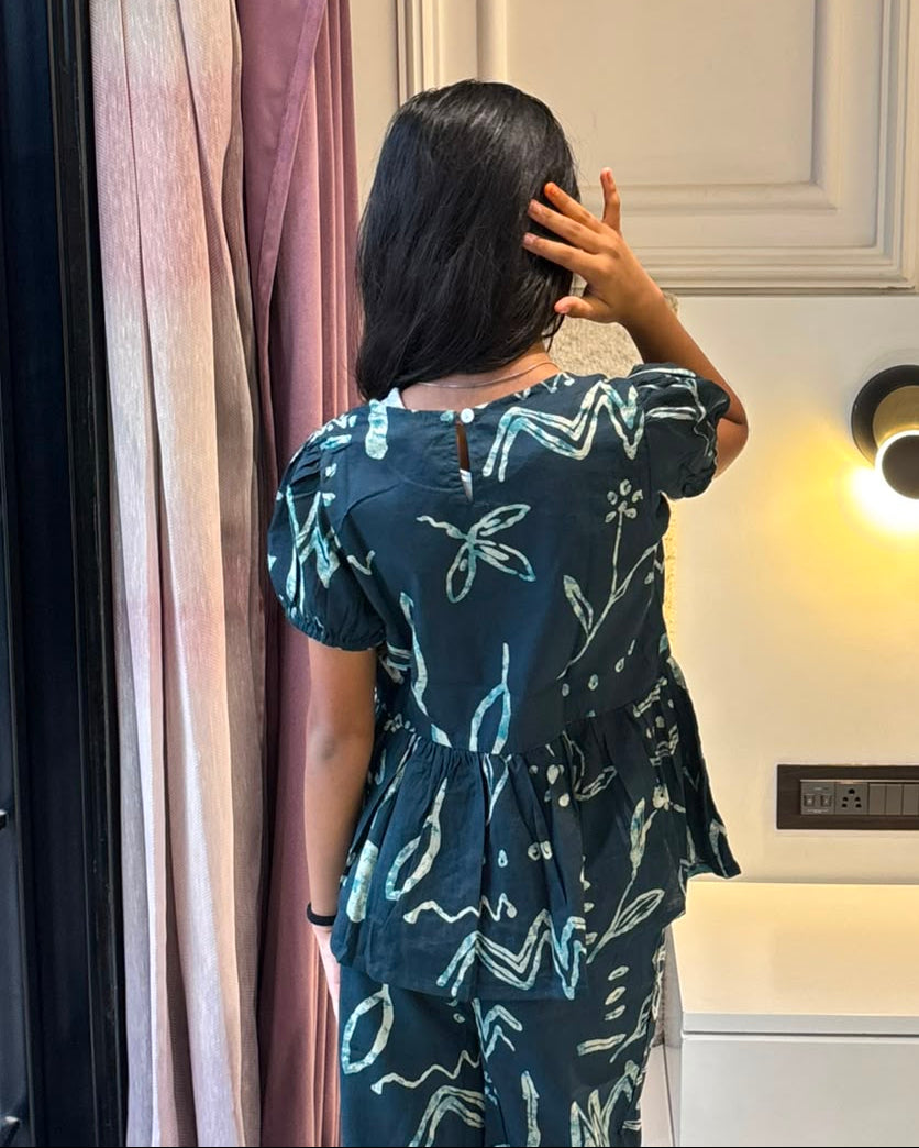 A girl is wearing a peplum styled top with matching pant in navy blue color from brand Aruvo, standing near a glass door smiling