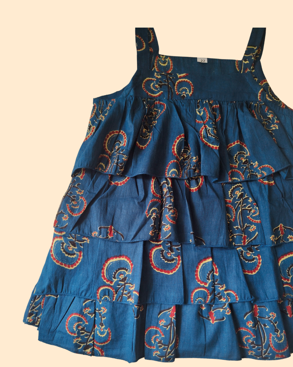 A tiered blue cotton dress with a red and gold paisley print, featuring thin shoulder straps and a ruffled skirt, shown against a solid beige background.