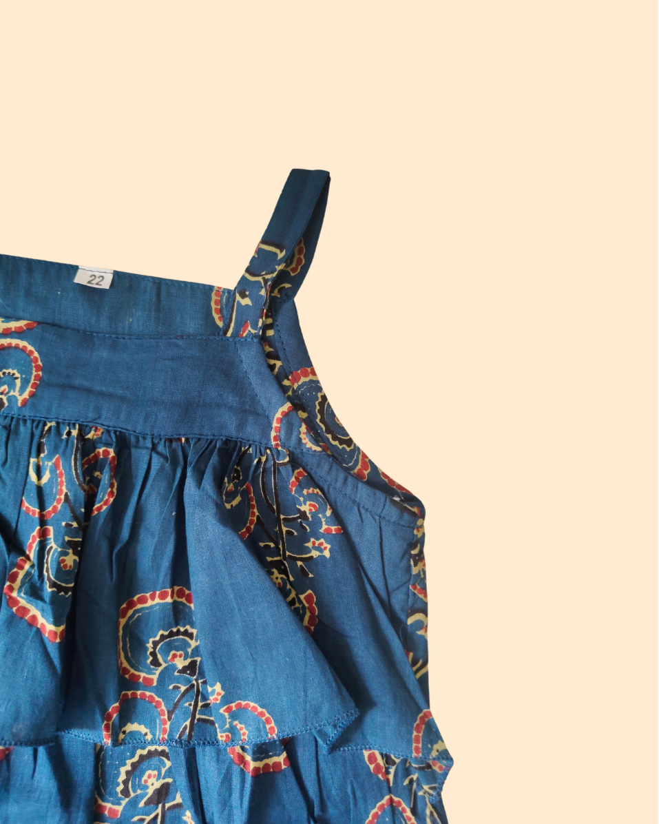 A close-up of a blue tiered cotton fabric garment, likely a ruffled top a red and gold paisley pattern and a single shoulder strap, set against a solid pale peach background.