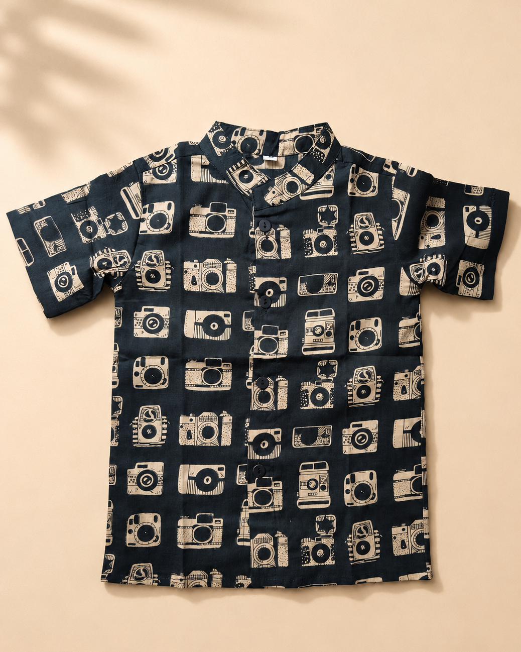 A short-sleeve button-up shirt with an all-over camera print, laid flat on a warm beige background.