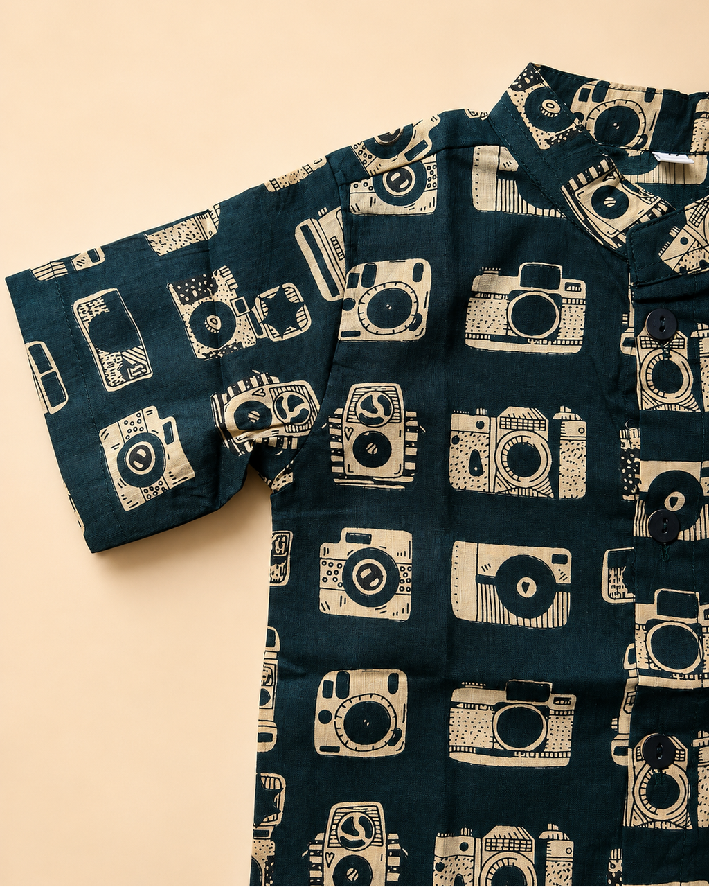 A short-sleeve button-up shirt with a dark teal background and a repeating cream-colored camera print, laid flat on a light beige background.