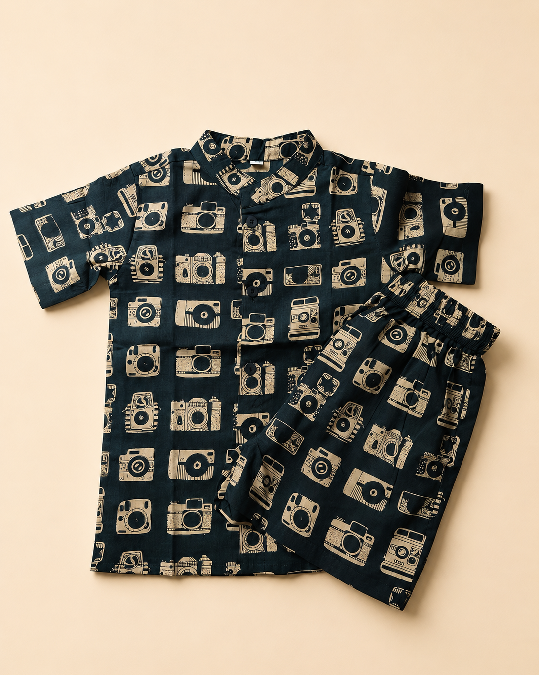 A two-piece children's pajama set featuring a short-sleeve button-up shirt and matching shorts, both printed with a repeating beige camera pattern on a dark navy blue background, laid flat on a warm beige background.