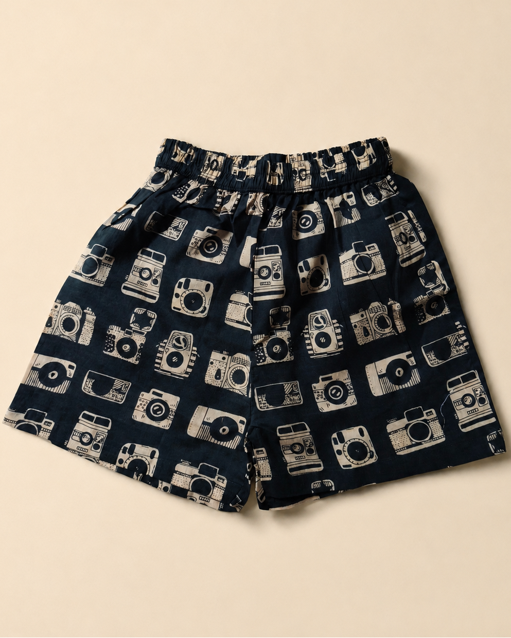 A pair of shorts with an elastic waistband, featuring a navy blue base with a repeating pattern of vintage film cameras in off-white and dark green, laid flat on a light beige background.
