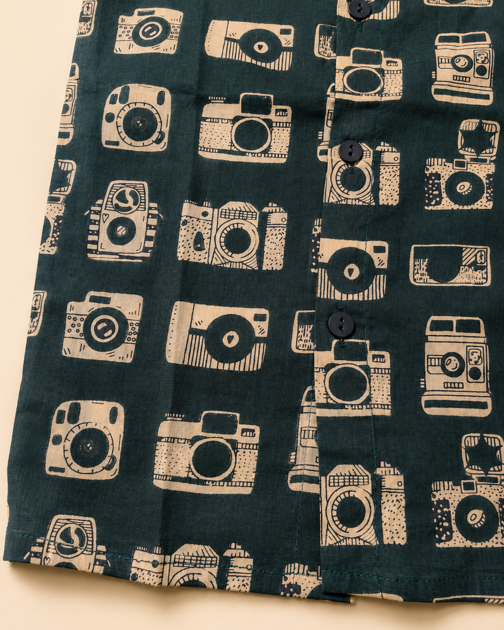 A close-up of a dark teal-green fabric with a repeating cream-colored vintage camera pattern, featuring a button-down front.