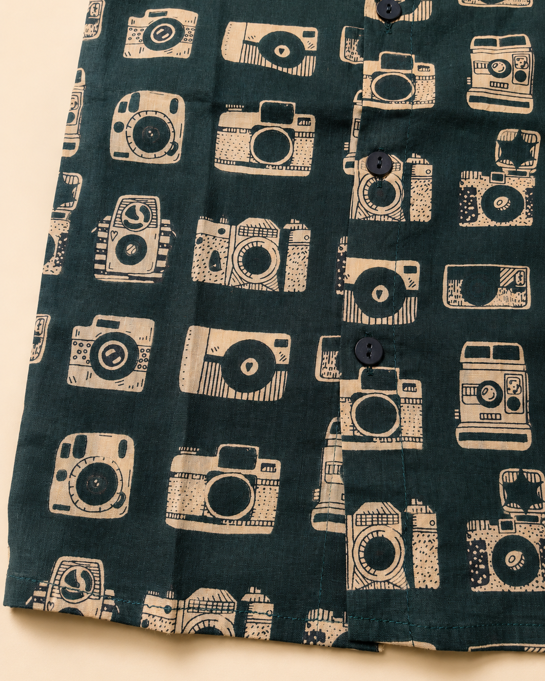 A close-up of a dark teal-green fabric with a repeating cream-colored vintage camera pattern, featuring a button-down front.