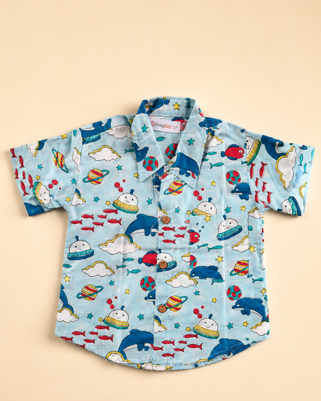 A light blue boys shirt with a colorful dolphin print, planets, rockets, clouds, and stars from Aruvo laid flat on a solid beige background.