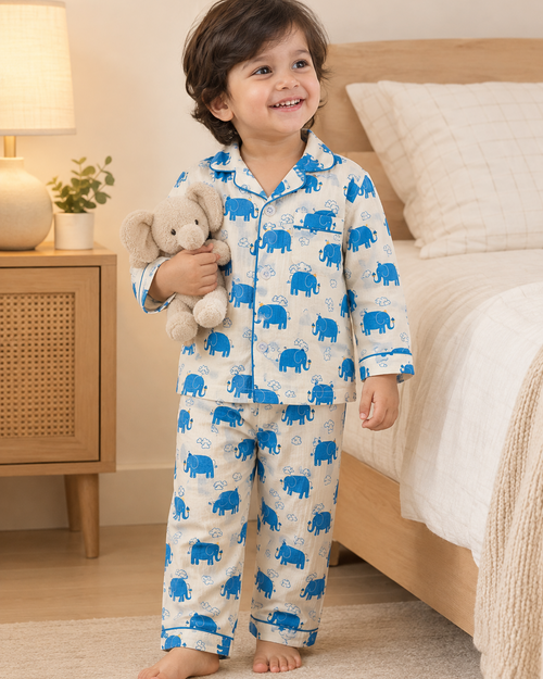 A smiling young child wearing a white pajama set with a blue elephant print stands barefoot in a bedroom, holding a plush elephant toy.