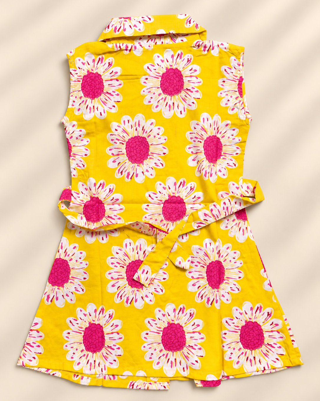 A yellow sleeveless dress with a large pink and white daisy print, featuring a collar, waist tie, and matching skirt, laid flat on a beige background with soft diagonal shadows.