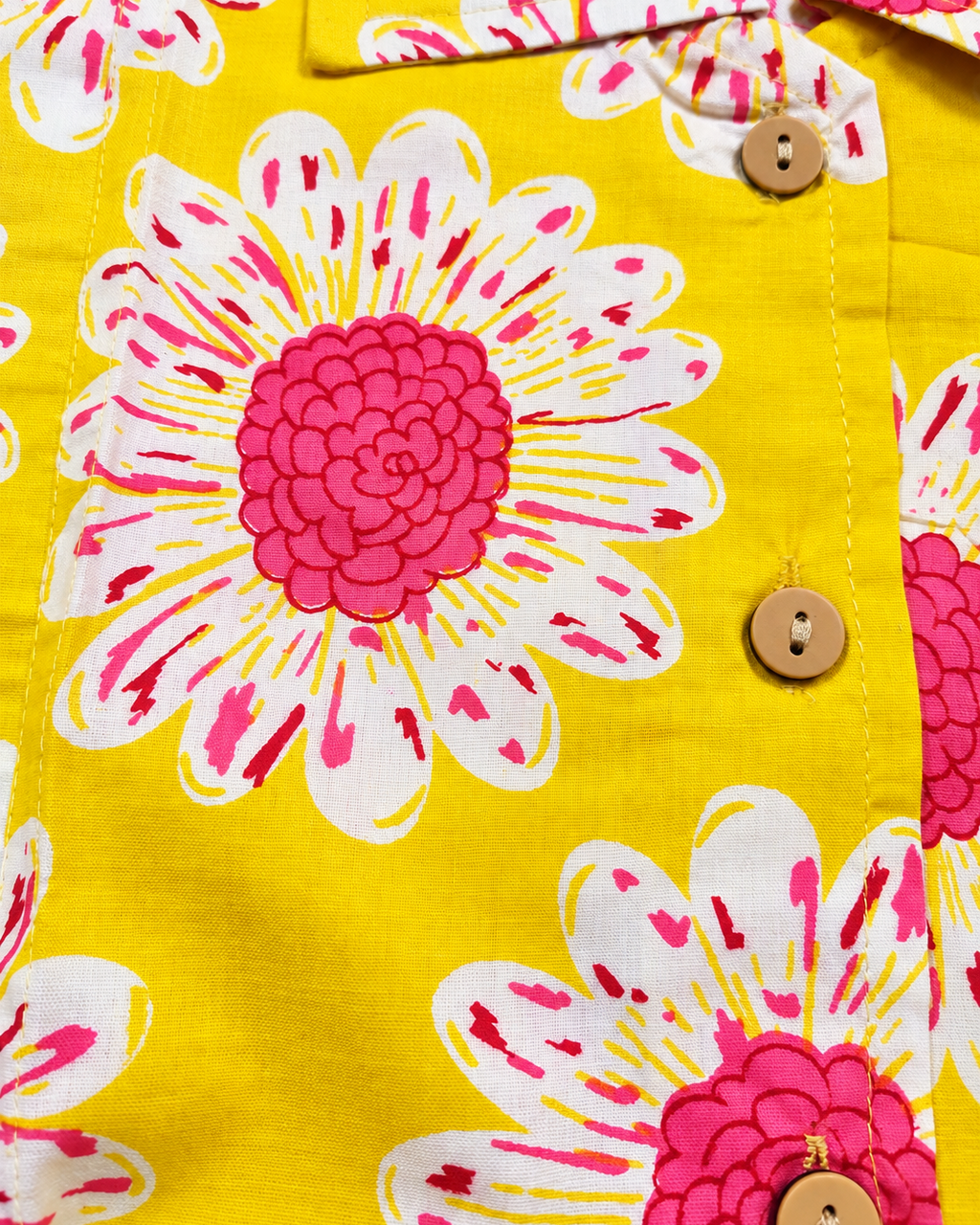 Close-up of a bright yellow button-front garment with a bold floral print featuring large white petals and vivid pink centers, plus three visible wooden buttons.