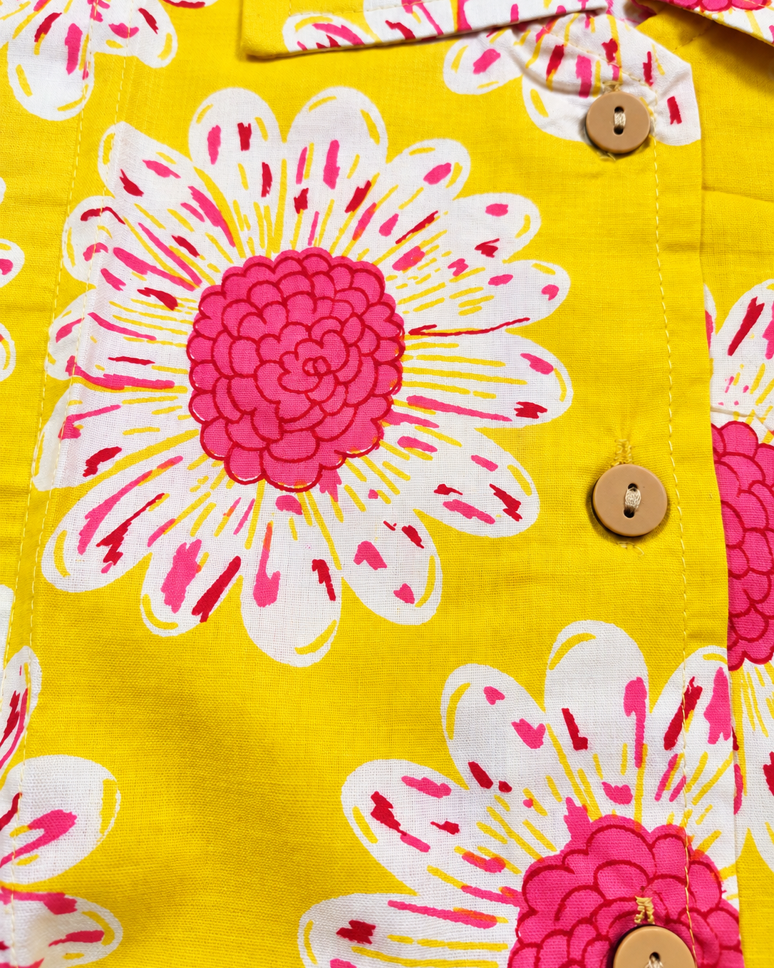 Close-up of a bright yellow button-front garment with a bold floral print featuring large white petals and vivid pink centers, plus three visible wooden buttons.