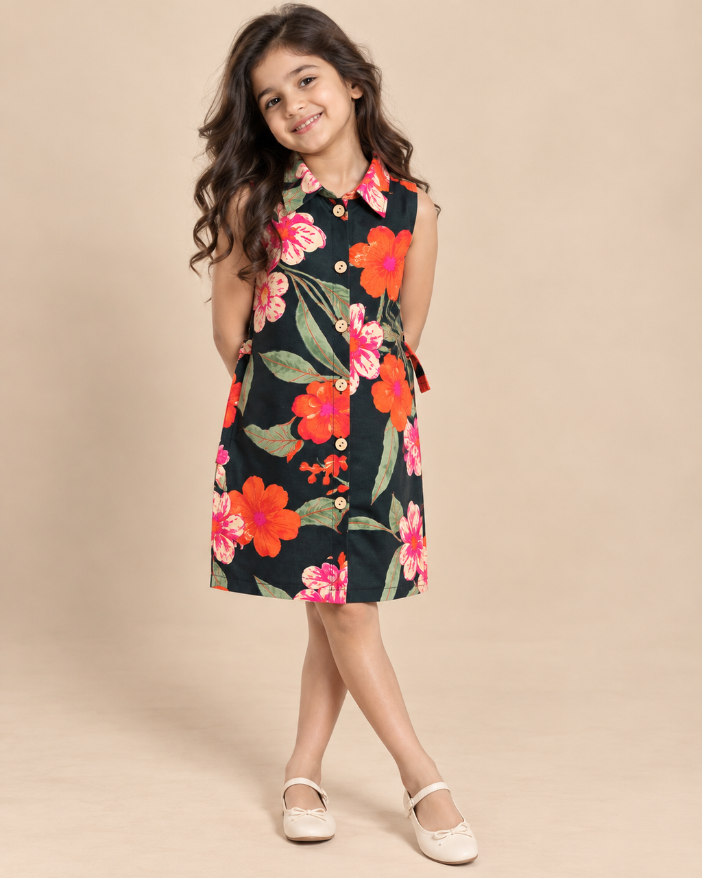 A young girl with long wavy brown hair stands against a solid beige studio background wearing a sleeveless dark green dress with a vibrant red, orange, and pink floral print, buttoned down the front, from brand Aruvo