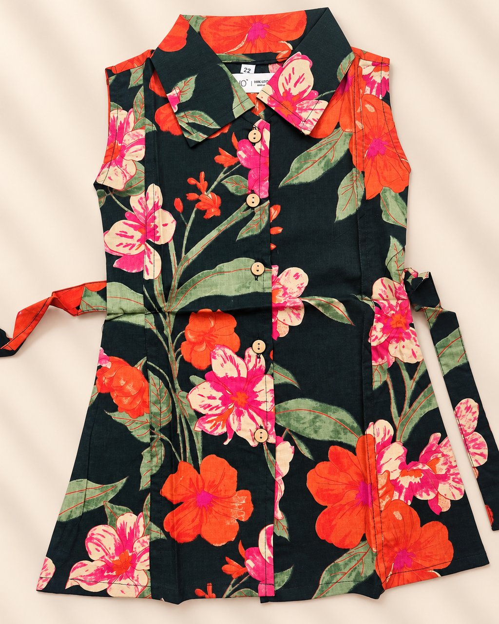 A floral apron laid flat on a light beige background with soft diagonal shadows. The apron features a black base with a vibrant pattern of pink, red, and orange flowers with green leaves.