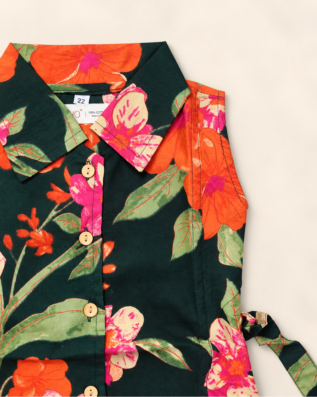 A close-up of a dark green floral shirt with orange, pink, and green hibiscus flowers, featuring a button-down front and laid flat on a light beige background.