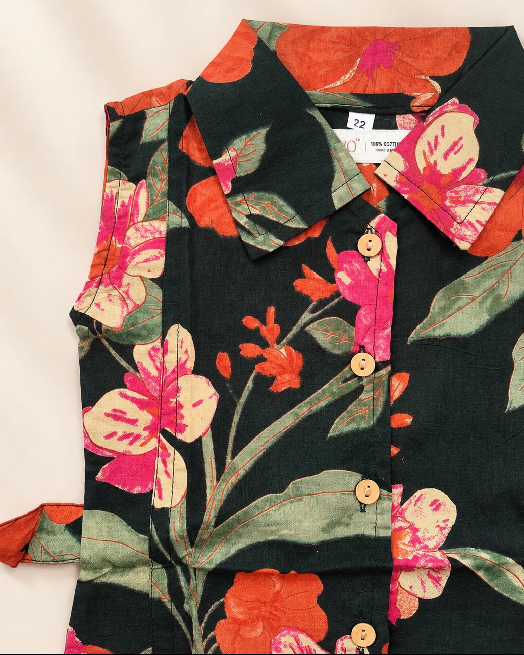 A close-up of a dark green floral shirt with orange, pink, and green hibiscus flowers, featuring a button-down front and a side tie detail, laid flat on a light beige background.