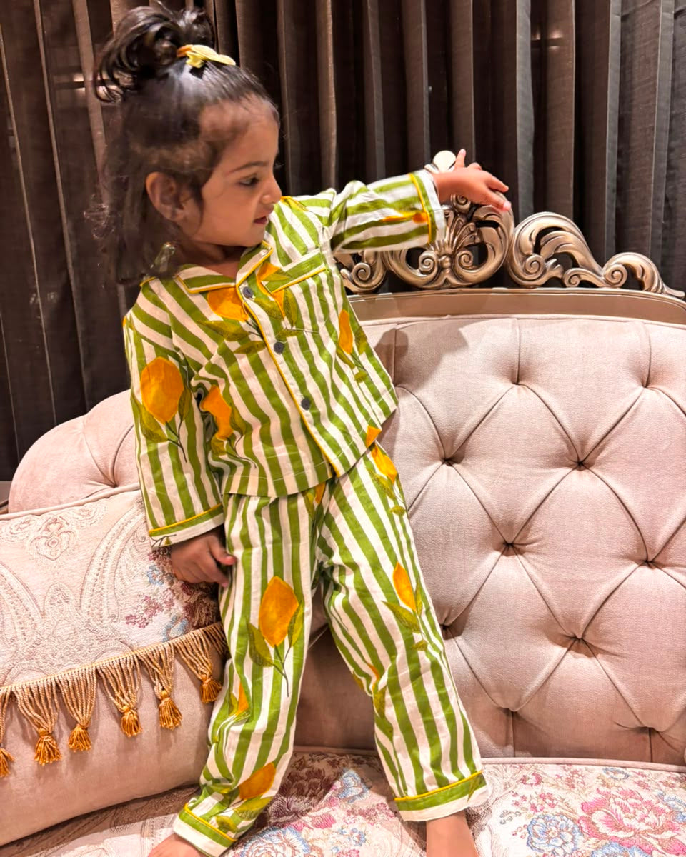A young girl is wearing a striped green and beige color full sleeve nightsuit set from brand Aruvo, standing on a bed smiling 