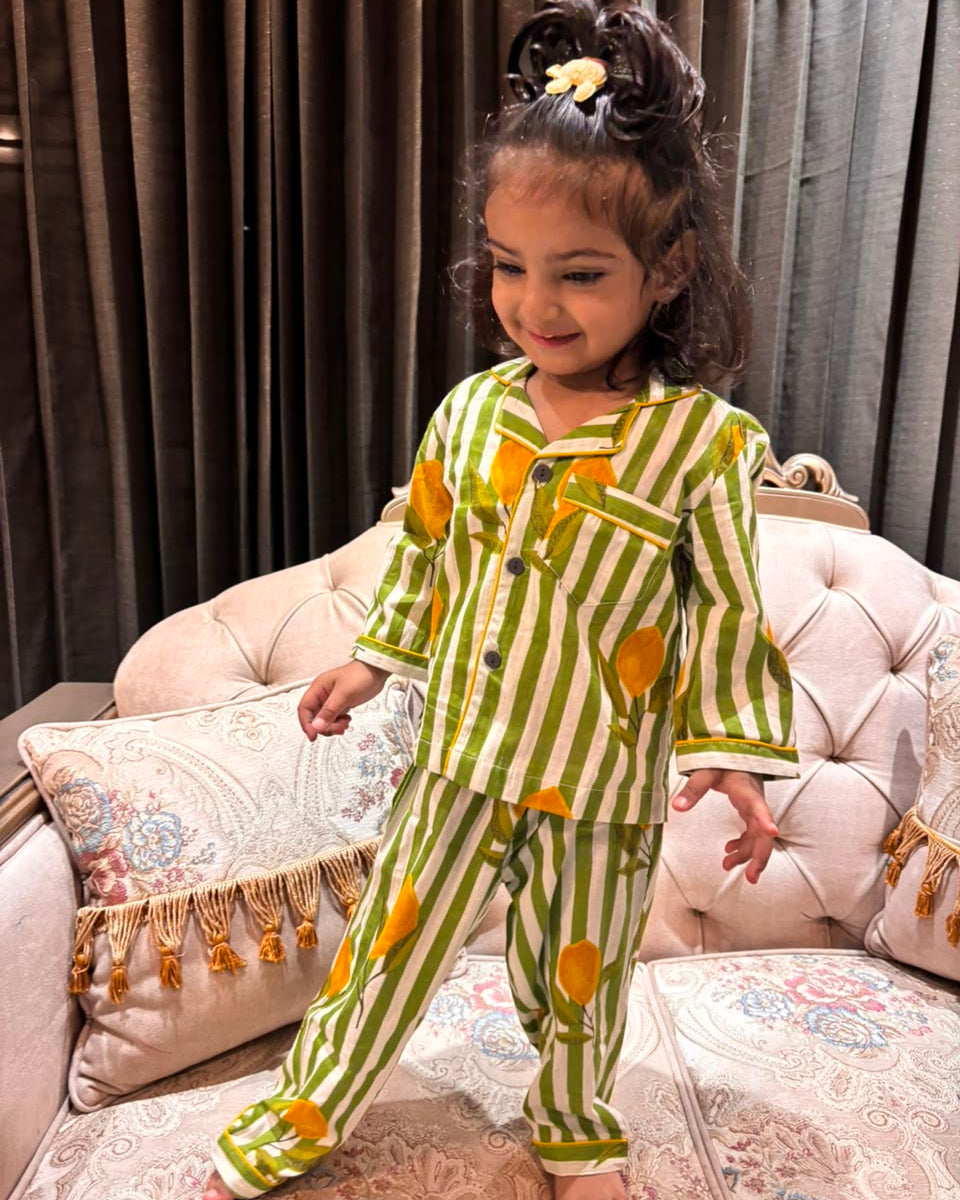 A young girl is wearing a striped green and beige color full sleeve nightsuit set from brand Aruvo, standing on a bed smiling 