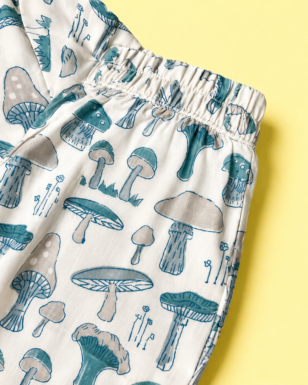 Close-up of a pair of white elastic-waist pants with a blue and gray mushroom print, laid flat on a solid yellow background.