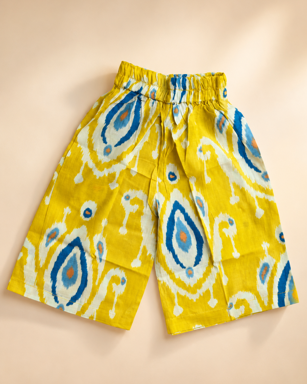 A pair of yellow cotton pant with a blue, white, and orange ikat pattern, featuring an elastic waistband, laid flat on a beige background.