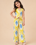 A young girl modeling a yellow sleeveless vest and matching wide-leg pants set with a blue and white ikat pattern from Aruvo, standing against a solid beige studio background.