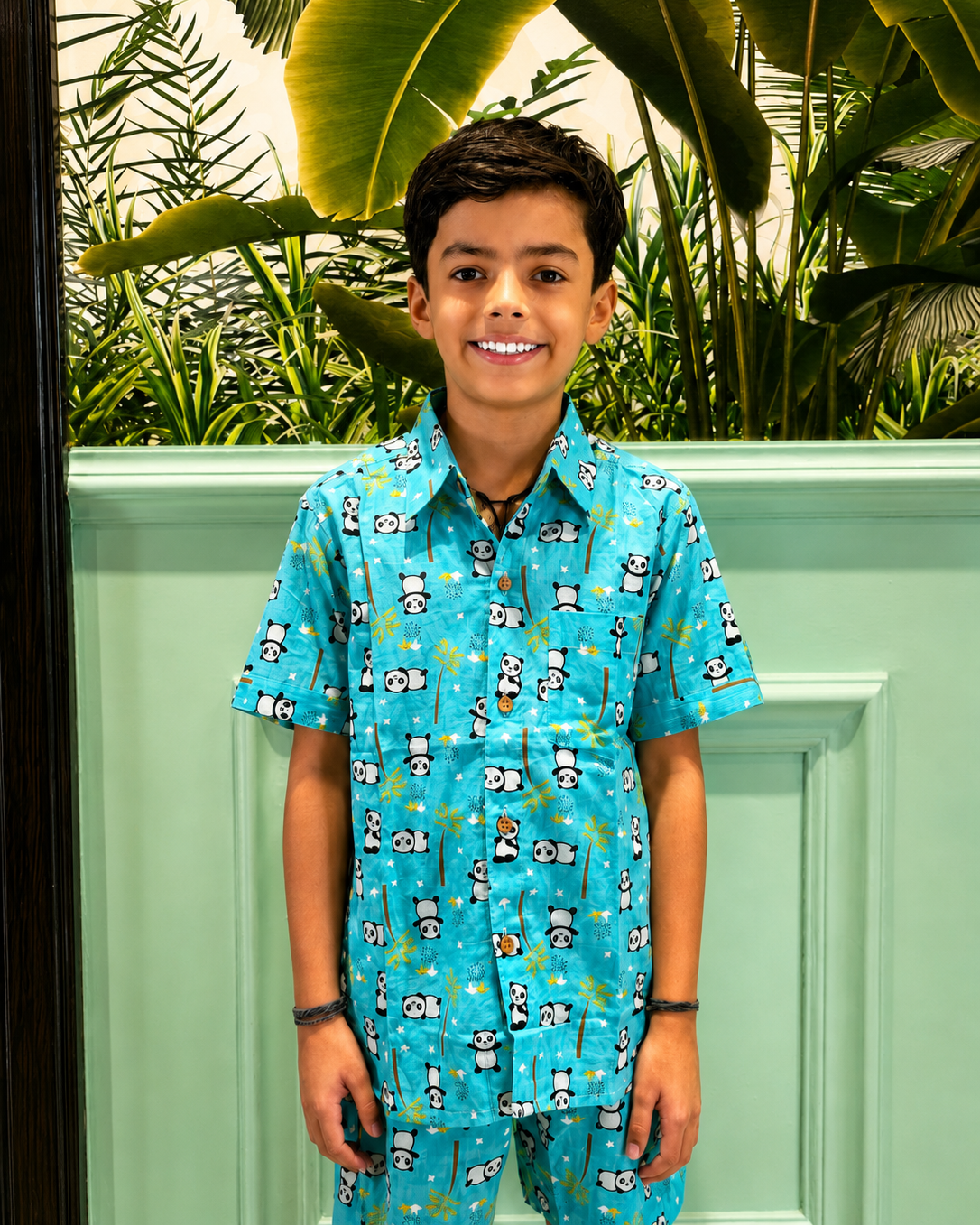 Child wearing Aruvo's blue patterned outfit, Panda prints on it standing in front of a textured wall with a plant.