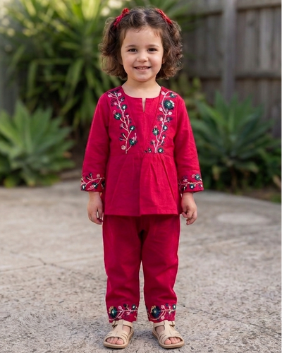 A young child wearing a bright pink embroidered top and matching pants from Aruvo, standing outdoors smiling