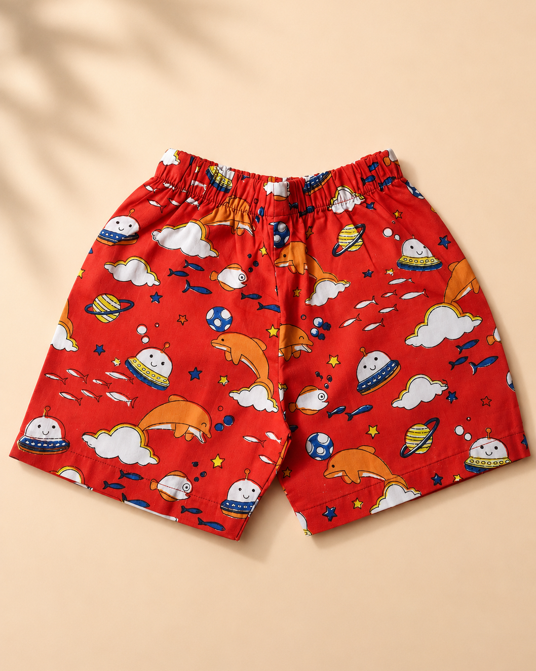 Red kids' shorts with a playful space-themed print featuring dolphins, planets, clouds, stars, and fish, displayed flat against a warm beige background.