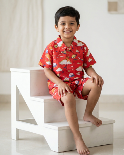 A young boy wearing a red short-sleeve button-up shirt and matching shorts with a playful dolphin, cloud and rocket print from Aruvo, sitting on white wooden stairs in a bright indoor setting.