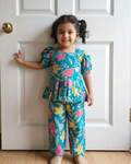 Child wearing a colorful floral outfit from Aruvo, standing in front of a white door.
