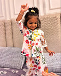 A toddler is wearing a loral printed full sleeve nightsuit set in white color from brand Aruvo, standing on a bed smiling 