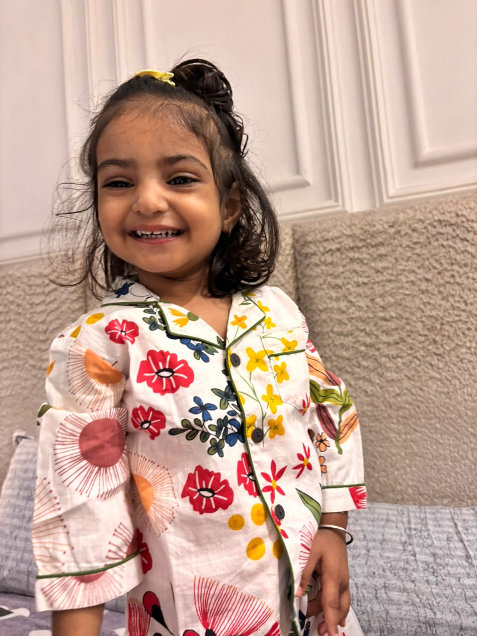 A toddler is wearing a loral printed full sleeve nightsuit set in white color from brand Aruvo, standing on a bed smiling 
