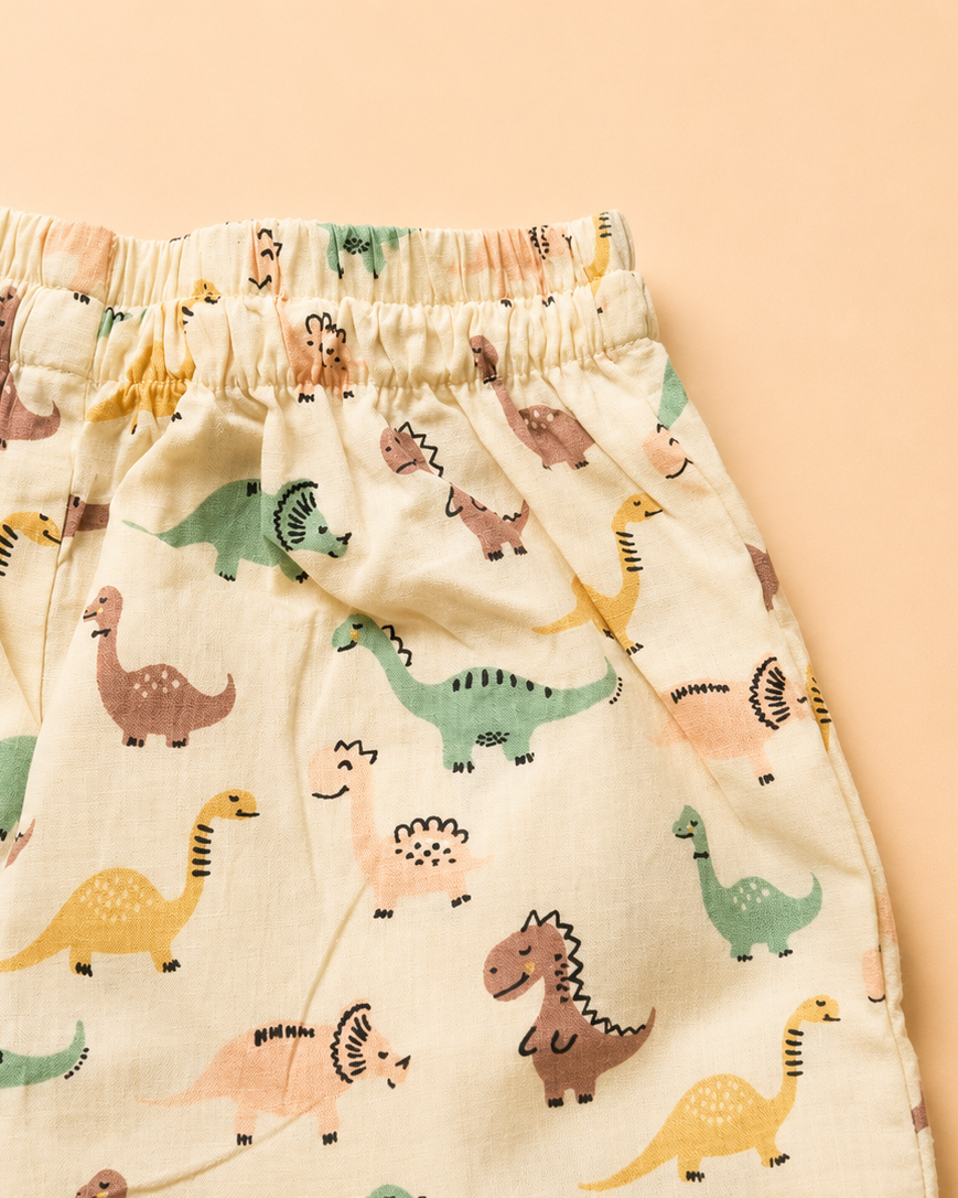 A close-up of a pair of children's shorts featuring an elastic waistband and an all-over dinosaur print on a cream-colored fabric, set against a solid peach background.