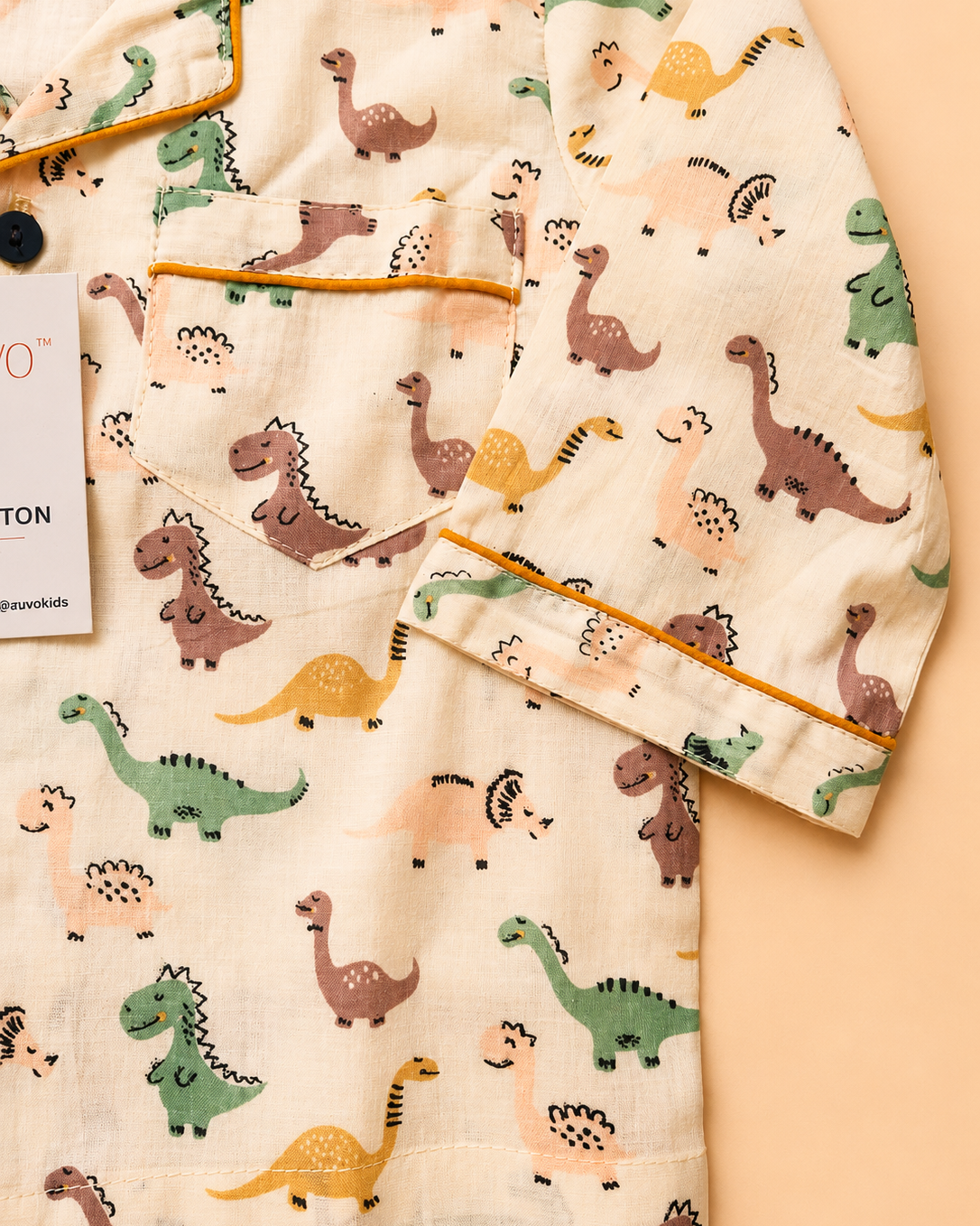 A close-up of a children's pajama top featuring an all-over dinosaur print on a cream-colored fabric, with mustard yellow piping along the collar, pocket, and cuffs from Aruvo