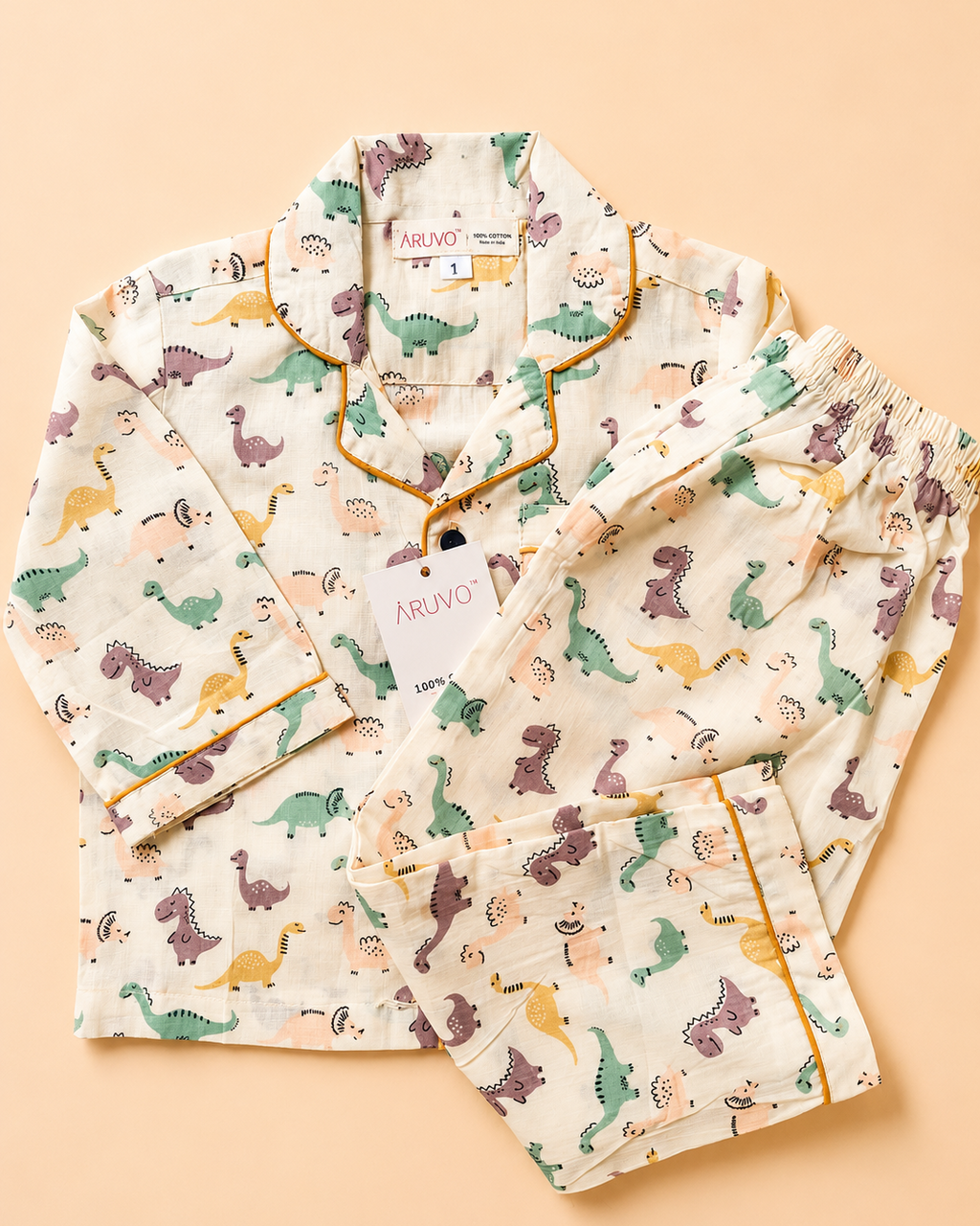 A two-piece children's pajama set laid flat on a warm peach background. The set includes a long-sleeve top and matching shorts in an off-white fabric printed with a colorful dinosaur pattern, finished with yellow piping along the collar, cuffs, and pocket edges.