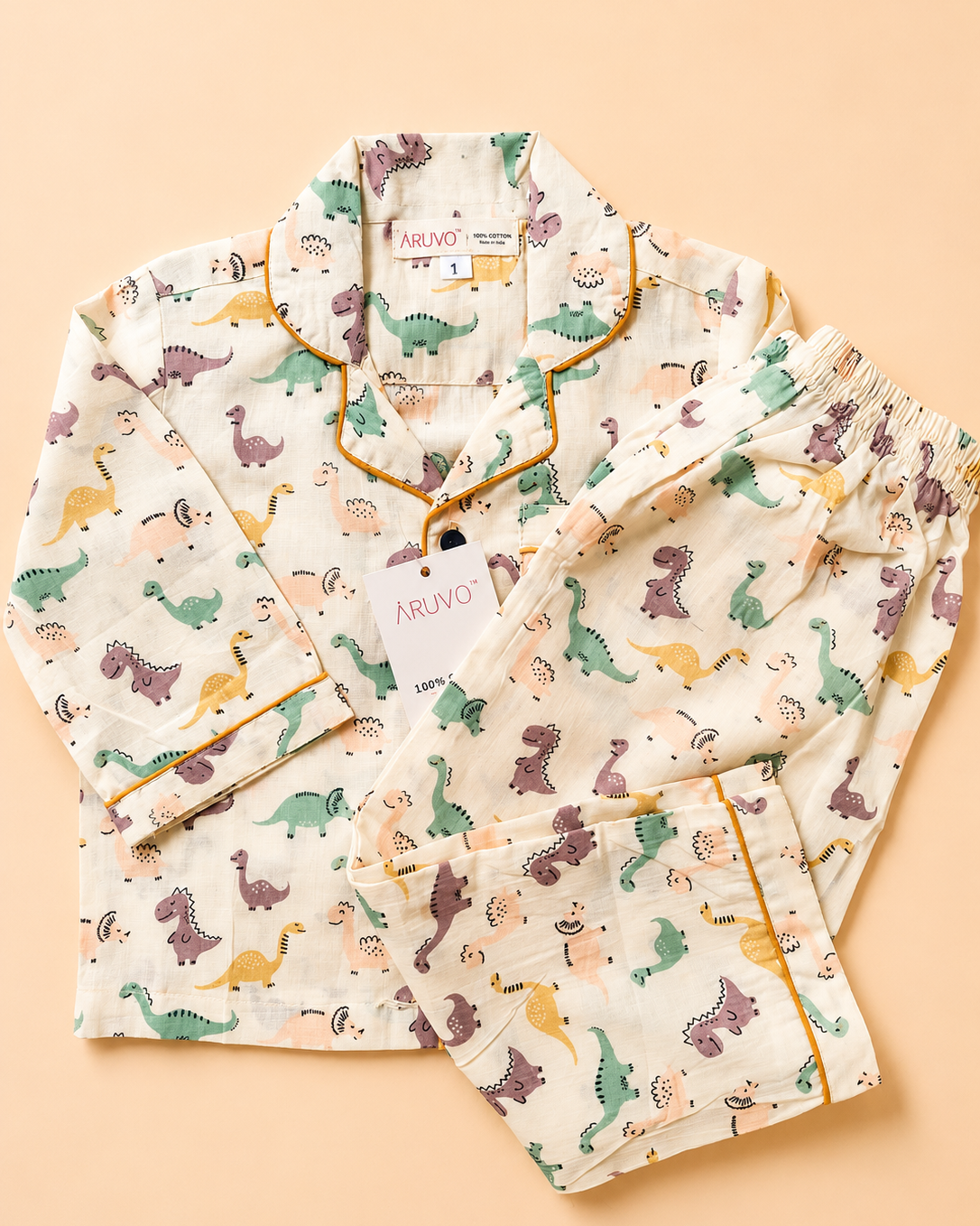 A two-piece children's pajama set laid flat on a warm peach background. The set includes a long-sleeve top and matching shorts in an off-white fabric printed with a colorful dinosaur pattern, finished with yellow piping along the collar, cuffs, and pocket edges.