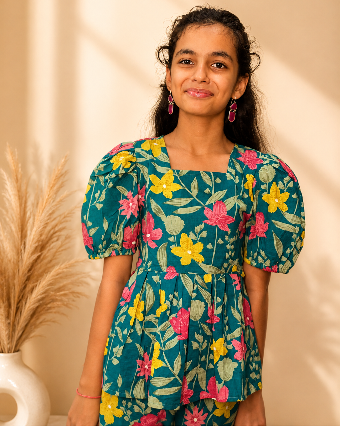 A young girl with dark hair tied back wears a teal puff-sleeve dress with a colorful floral print of pink, yellow, and green flowers. She is standing in front of a warm beige wall with soft shadows, next to a white vase with dried pampas grass.